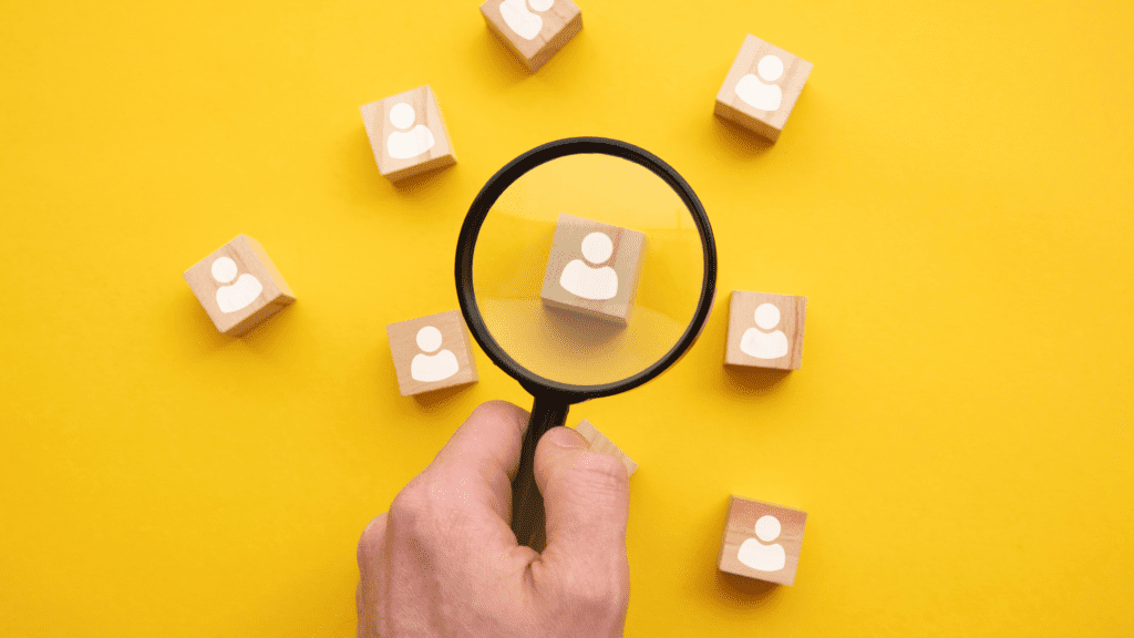 Scattered wooden blocks with white icons symbolizing a salesperson on each block with a magnifying glass focused on one of the blocks - depicting How to Hire Great Salespeople